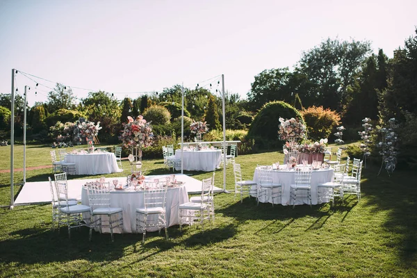 Banquet table on a green lawn, dance floor. Racks and cutlery, velvet napkins, pink glasses. Floral arrangement of pink flowers. Silver candlesticks and candles. On the table is a white tablecloth.