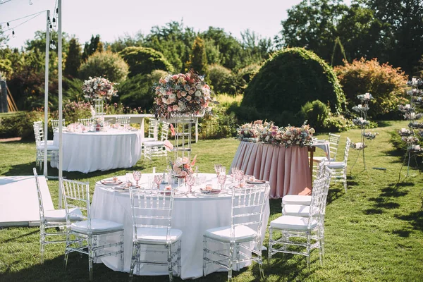 Banquet table on a green lawn, dance floor. Racks and cutlery, velvet napkins, pink glasses. Floral arrangement of pink flowers. Silver candlesticks and candles. On the table is a white tablecloth.