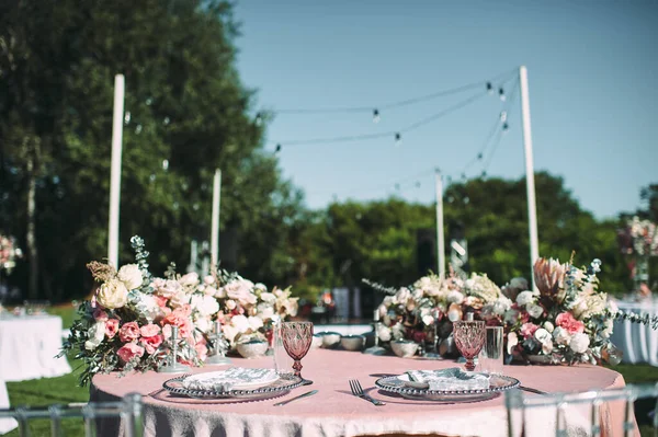 Banquet table on a green lawn. Racks and cutlery, velvet napkins, pink glasses. Floral arrangement of pink flowers. Silver candlesticks and candles. On the table is a white tablecloth.