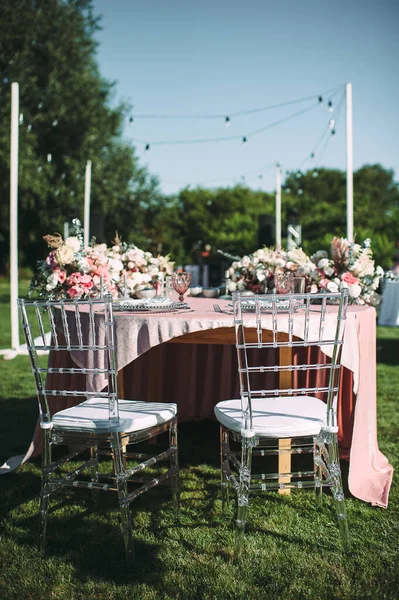 Banquet table on a green lawn. Racks and cutlery, velvet napkins, pink glasses. Floral arrangement of pink flowers. Silver candlesticks and candles. On the table is a white tablecloth.
