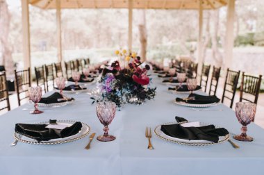 Doğadaki ziyafet masası. Çatal bıçak takımı, mor gözlükler, siyah peçeteler. Menekşe, mavi, siyah, gümüş çiçekler, anturyum, güller, karanfiller, östomalar. Mavi masa örtüsü.