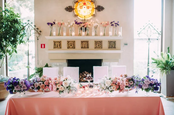 Presidium, banquet table with a pink tablecloth on the background of the fireplace in a medieval style. Plates and cutlery, purple glasses, napkins, candles in glass candlesticks. Bouquet of flowers.