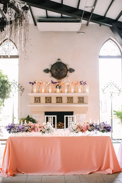 Presidium, banquet table with a pink tablecloth on the background of the fireplace in a medieval style. Plates and cutlery, purple glasses, napkins, candles in glass candlesticks. Bouquet of flowers.