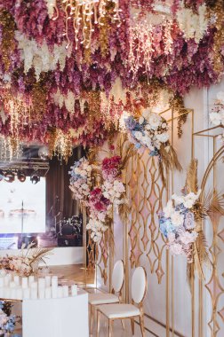 Beyaz ziyafet masası, başkanlık kürsüsü, mumlar, pembe, mavi, leylak, beyaz, gül, ortanca, altın yapraklar, salkım. Çiçekli altın rafların arkasında.