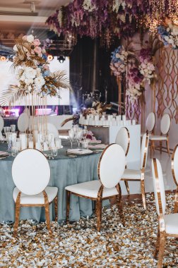 Mavi kadife masa örtülü ziyafet masası, mumlar, Alexa Yemek Sandalyesi. Gül çiçekleri, ortancalar ve altın yapraklardan oluşan yüksek bir tabaka. Altın rafların arkasında çiçekler, sahne, ekran, ışık.