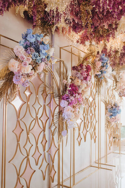 Banquet white table, presidium, candles, composition of flowers of pink, blue, lilac, white, rose, hydrangea, golden leaves, wisteria. Against the background of golden racks with flowers.