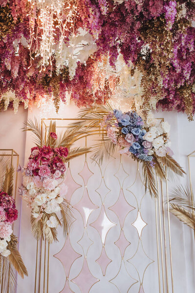 Banquet white table, presidium, candles, composition of flowers of pink, blue, lilac, white, rose, hydrangea, golden leaves, wisteria. Against the background of golden racks with flowers.