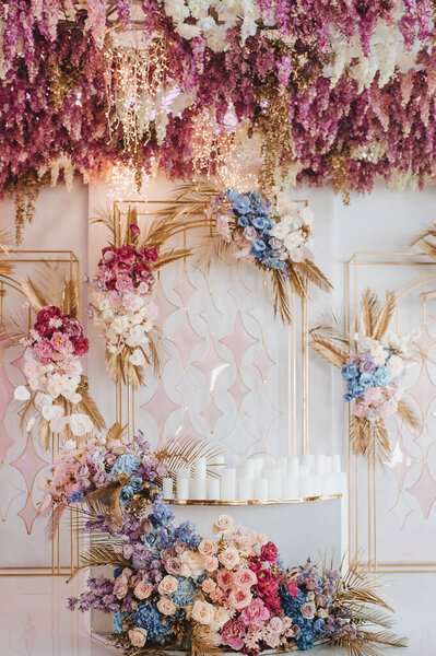 Banquet white table, presidium, candles, composition of flowers of pink, blue, lilac, white, rose, hydrangea, golden leaves, wisteria. Against the background of golden racks with flowers.