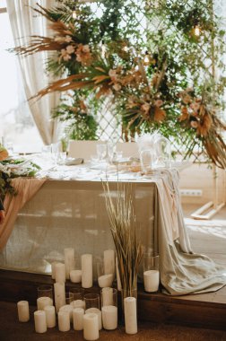 Yeni evlilerin masası, başkanlık, gri keten bir masa örtüsü, çiçeklerden oluşan bir kompozisyon, arka planda tropikal yapraklar ve ahududu dallarıyla süslenmiş bir kafes.