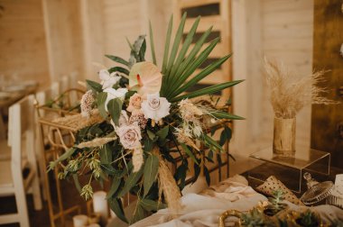 Restoran, servis edilen masa, köy yemekleri, büyük çiçek kompozisyonları ve masada tropikal yapraklar, tabaklar, çatal bıçak takımı, kristal bardaklar, ahşap sandalyeler, ampul çelenkleri..