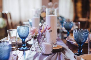 Taşralı. Masa beyaz bir masa örtüsüyle kaplı, servis ediliyor. Tabaklar, çatal bıçak, peçete, mavi bardaklar, oturma kartları, yeşillikler, cam vazodaki mumlar, mavi ışık, kurumuş çiçekler..