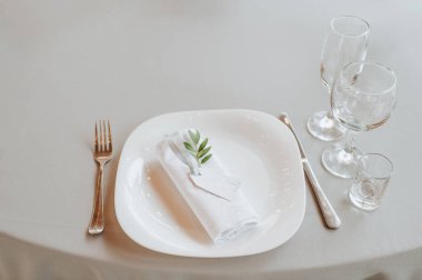 Masa beyaz bir masa örtüsüyle kaplı, servis ediliyor. Tabaklar, çatal bıçak, peçete, mavi bardaklar, oturma kartları, yeşillik filizi.