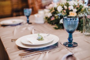 Masa beyaz bir masa örtüsüyle kaplı, servis ediliyor. Tabaklar, çatal bıçak, peçete, mavi bardaklar, oturma kartları, yeşillik filizi.
