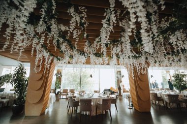 Modern ahşap arka plan, çelenk, iç mekan. Masalar servis edildi, çiçekler ve yeşillik kompozisyonları. Tabaklar, çatal bıçak takımı, peçete, pembe bardaklar. Yemek salonu..