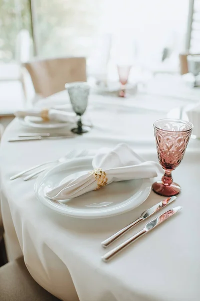 The table is covered with a white tablecloth, served. Plates, cutlery, napkin, pink glasses, seating cards, sprig of greens.