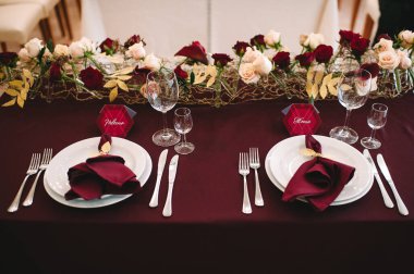 Marsala masa örtülü ziyafet masası, mumlar, set kartları, peçeteler, tabaklar, çatal bıçak takımı, bardaklar, marsala gülünden çiçekler. Başlık: 