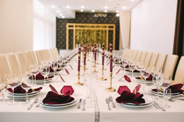 Banquet table with white tablecloth, candles, setting cards, napkins, plates, cutlery, glasses, composition of flowers of marsala rose. Candles, decor in gold, wine and marsala colors