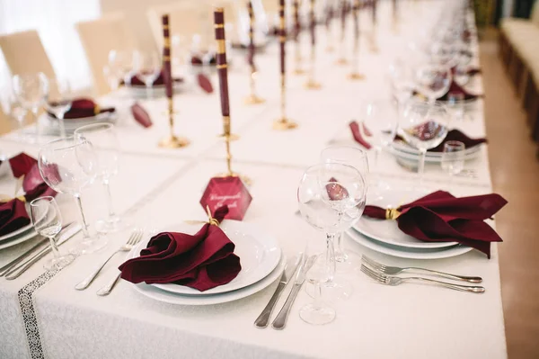 Banquet table with white tablecloth, candles, setting cards, napkins, plates, cutlery, glasses, composition of flowers of marsala rose. Candles, decor in gold, wine and marsala colors