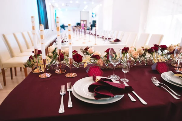 Banquet table with marsala tablecloth, candles, setting cards, napkins, plates, cutlery, glasses, composition of flowers of marsala rose. Candles, decor in gold, wine and marsala colors
