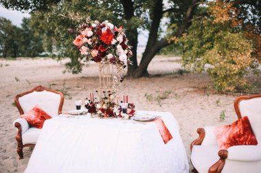 Doğayla ilgili bir ziyafet, beyaz masa örtülü bir masa, altın kenarlı klasik tabaklar, şarap bardakları, çatal bıçak takımı, mumlar, çiçeklerden oluşan bir kompozisyon, amirilis, güller, kırmızı yıldız çiçekleri..