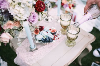 Kurabiyeler, gümüş bir vazoda incir, bir buket gül ve bir yıldız çiçeği, masa örtüsüyle kaplı yeşil bardaklar. Mavi koltuklar. Yeşil çimlerin ve ağaçların arka planında