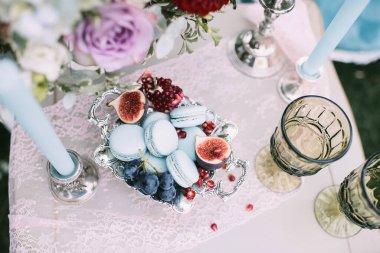 Kurabiyeler, gümüş bir vazoda incir, bir buket gül ve bir yıldız çiçeği, masa örtüsüyle kaplı yeşil bardaklar. Mavi koltuklar. Yeşil çimlerin ve ağaçların arka planında