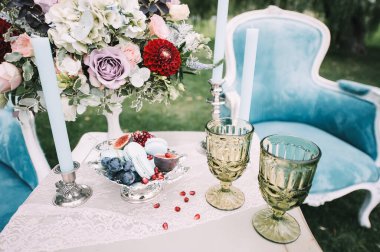 Kurabiyeler, gümüş bir vazoda incir, bir buket gül ve bir yıldız çiçeği, masa örtüsüyle kaplı yeşil bardaklar. Mavi koltuklar. Yeşil çimlerin ve ağaçların arka planında