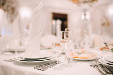 Ziyafet, restoran. Masa ayarı. Beyaz tabak, gözlük? Çatal bıçak. Masanın üzerinde beyaz bir masa örtüsü, peçete ve yiyecek var. Açık renkli çiçek aranjmanları.