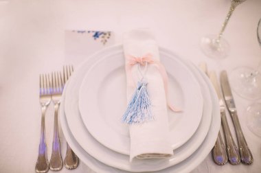 Ziyafet, restoran. Masa ayarı. Beyaz tabak, gözlük? Çatal bıçak. Masanın üzerinde beyaz bir masa örtüsü, peçete ve yiyecek var. Açık renkli çiçek aranjmanları.