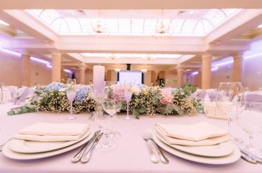 Ziyafet, restoran. Masa ayarı. Beyaz tabak, gözlük? Çatal bıçak. Masanın üzerinde beyaz bir masa örtüsü, peçete ve yiyecek var. Açık renkli çiçek aranjmanları.