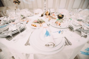 Ziyafet, restoran. Masa ayarı. Beyaz tabak, gözlük? Çatal bıçak. Masanın üzerinde beyaz bir masa örtüsü, peçete ve yiyecek var. Açık renkli çiçek aranjmanları.