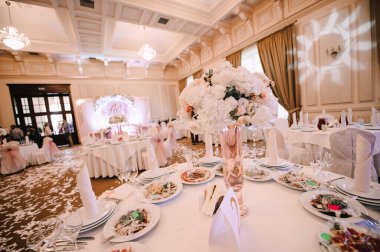 Ziyafet, restoran. Masa ayarı. Beyaz tabak, gözlük? Çatal bıçak. Masanın üzerinde beyaz bir masa örtüsü, peçete ve yiyecek var. Açık renkli çiçek aranjmanları.