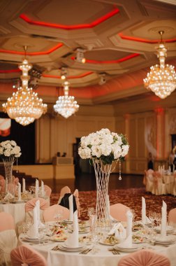 Restoran, ziyafet, sofra kurma. Masa beyaz bir masa örtüsü, bulaşıklar, beyaz temiz tabaklar, çatal bıçak takımı, peçeteler, şarap bardakları, mumlarla kaplı. Yakın plan. Büyük kristal avizeler.