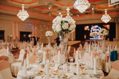 Restoran, ziyafet, sofra kurma. Masa beyaz bir masa örtüsü, bulaşıklar, beyaz temiz tabaklar, çatal bıçak takımı, peçeteler, şarap bardakları, mumlarla kaplı. Yakın plan. Büyük kristal avizeler.