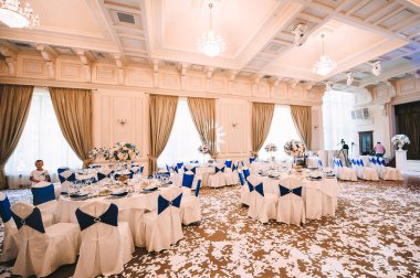 Ziyafet, restoran. Masa ayarı. Beyaz tabak, gözlük? Çatal bıçak. Masanın üzerinde beyaz bir masa örtüsü, peçete ve yiyecek var. Açık renkli çiçek aranjmanları.