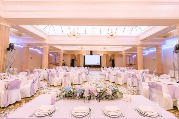 Ziyafet, restoran. Masa ayarı. Beyaz tabak, gözlük? Çatal bıçak. Masanın üzerinde beyaz bir masa örtüsü, peçete ve yiyecek var. Açık renkli çiçek aranjmanları.