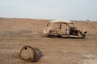Kızgın gökyüzü ve pas fıçısıyla çöl manzarasında hurda bir araba.