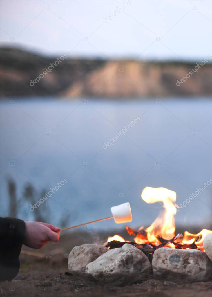 person roasting marshmallows over an open campfire in the late summer nights while camping by a lake blurred background for copy space and text overlay 