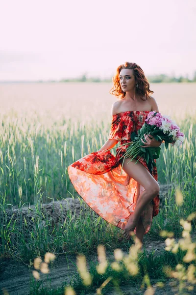 Redhead girl in a red dress with a bouquet of peonies dancing joyful in a wheat field in summer at sunset. Soft focus. Back light. Vertical