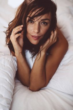 Sabahları beyaz bir yatakta uyanan güzel bir genç kız, yeni güne gülümsüyor ve pencereden güneşi görüyor ve yüzünü saçlarla örtüyor. Yumuşak seçici odaklanma. Renkli fotoğraf.