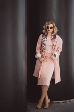 Zarif bir moda. Pembe elbiseli ve güneş gözlüklü şık sarışın kız siyah büyük fıçıların arkasında poz veriyor. Yumuşak seçici odak.