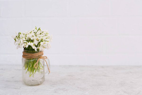 Bouquet of spring flowers of snowdrops in a glass vase on a light brick wall. Copy space for greeting card on mother's day.