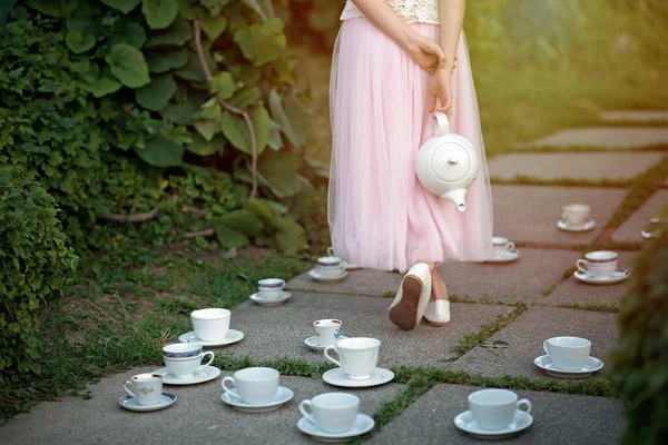 Alice in Wonderland. A girl in a full skirt with a teapot in her hands among a set of cups and saucers.