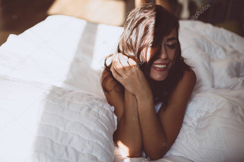 A beautiful young girl in the morning in a white bed after waking up, smiles at the new day and the sun from the window and covers her face with hair. Soft selective focus. Toned photo.