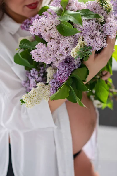Belly of a pregnant woman in a white shirt in the bathroom near the window with a bouquet of lilac flowers. Soft selective focus.