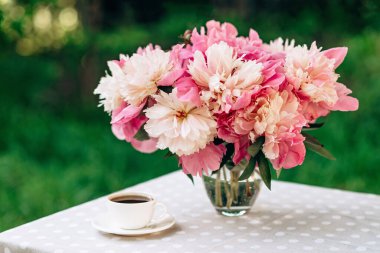 Açık havada samimi romantik bir kahvaltı. Şakayık çiçekleri vazosu, masada bir fincan kahve çayı. Yumuşak seçici odak.