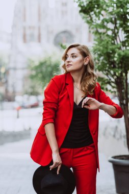 Kırmızı takım elbiseli ve siyah şapkalı güzel sarışın kadın binanın yanındaki avluda duruyor. Şık insanlar. Yumuşak seçici odak.