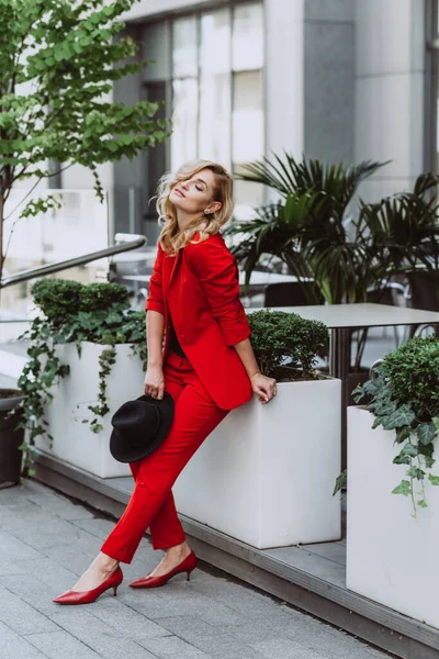 stock image Young beautiful blond woman in a red suit and black hat stands outdoors in the yard next to the building. Stylish people. Soft selective focus.