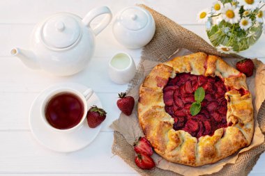 Taze pişmiş ev yapımı galet ya da açık çilekli turta, bir fincan bitki çayı, çaydanlık ve bir buket papatya çiçekli vazo. Yumuşak seçici odaklanma. Düz yatıyordu.