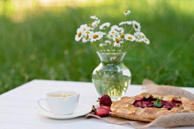 Taze pişmiş ev yapımı galet ya da açık çilekli turta, bir fincan bitki çayı ve bir buket papatya çiçekli vazo. Yumuşak seçici odak.